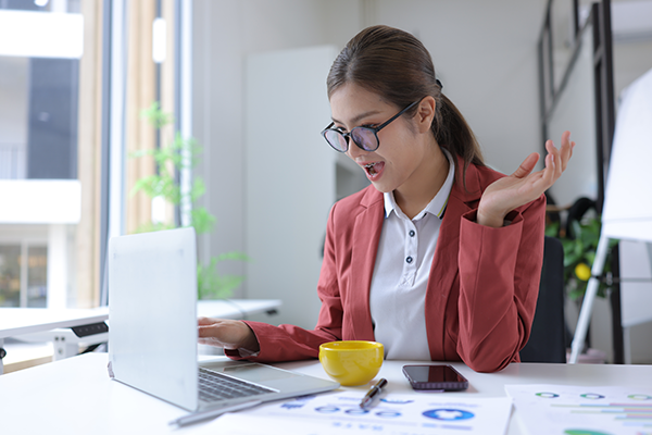 Professional woman excitedly reacting at laptop in office, with coffee cup and paperwork.