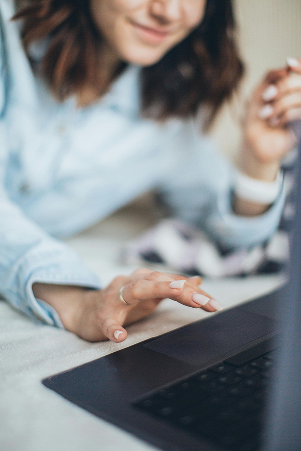 Woman using a laptop with focus on hand typing, wearing a light blue shirt, indoors, blurred background.