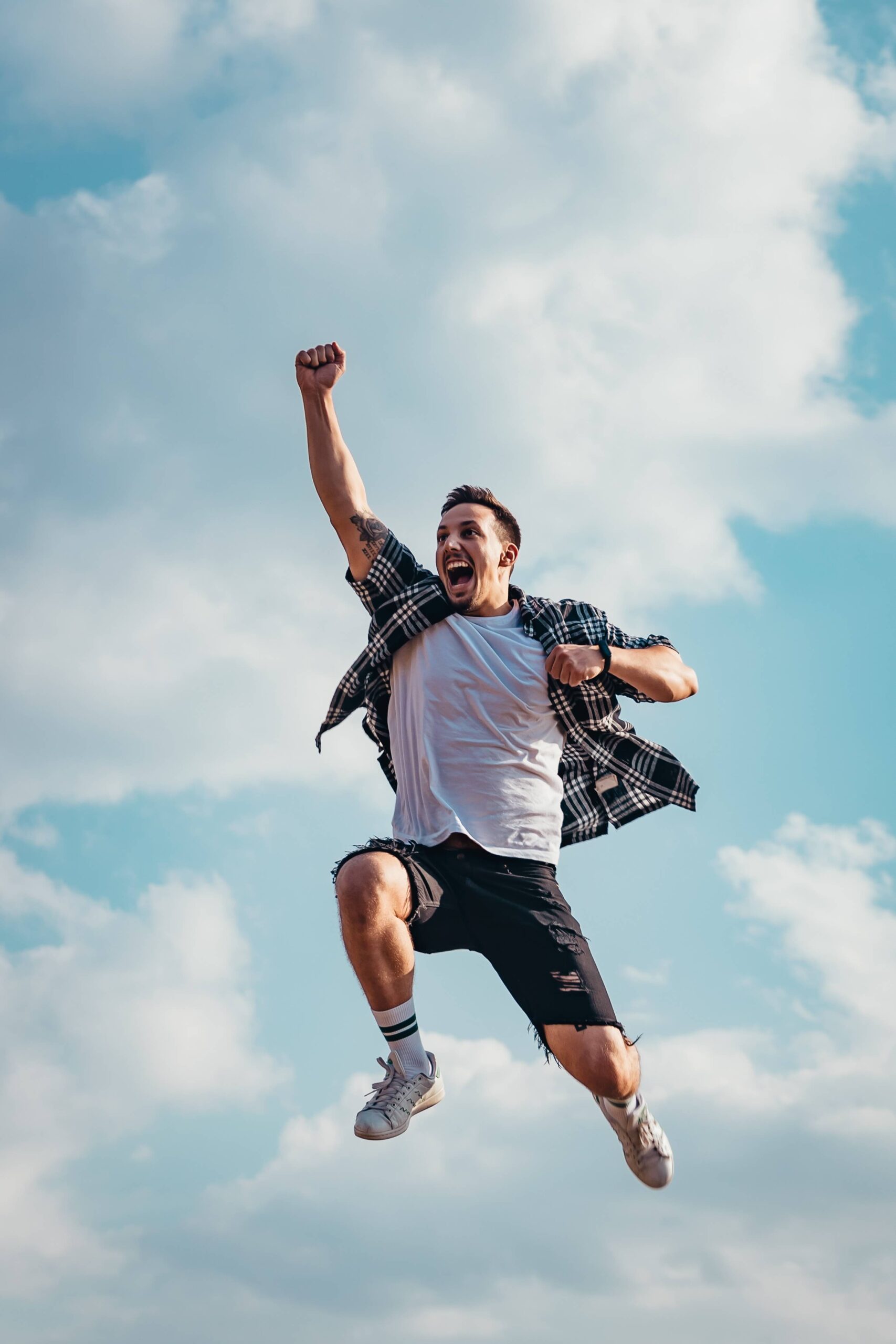 Man joyfully jumping against cloudy sky, wearing plaid shirt and shorts, expressing freedom and happiness.