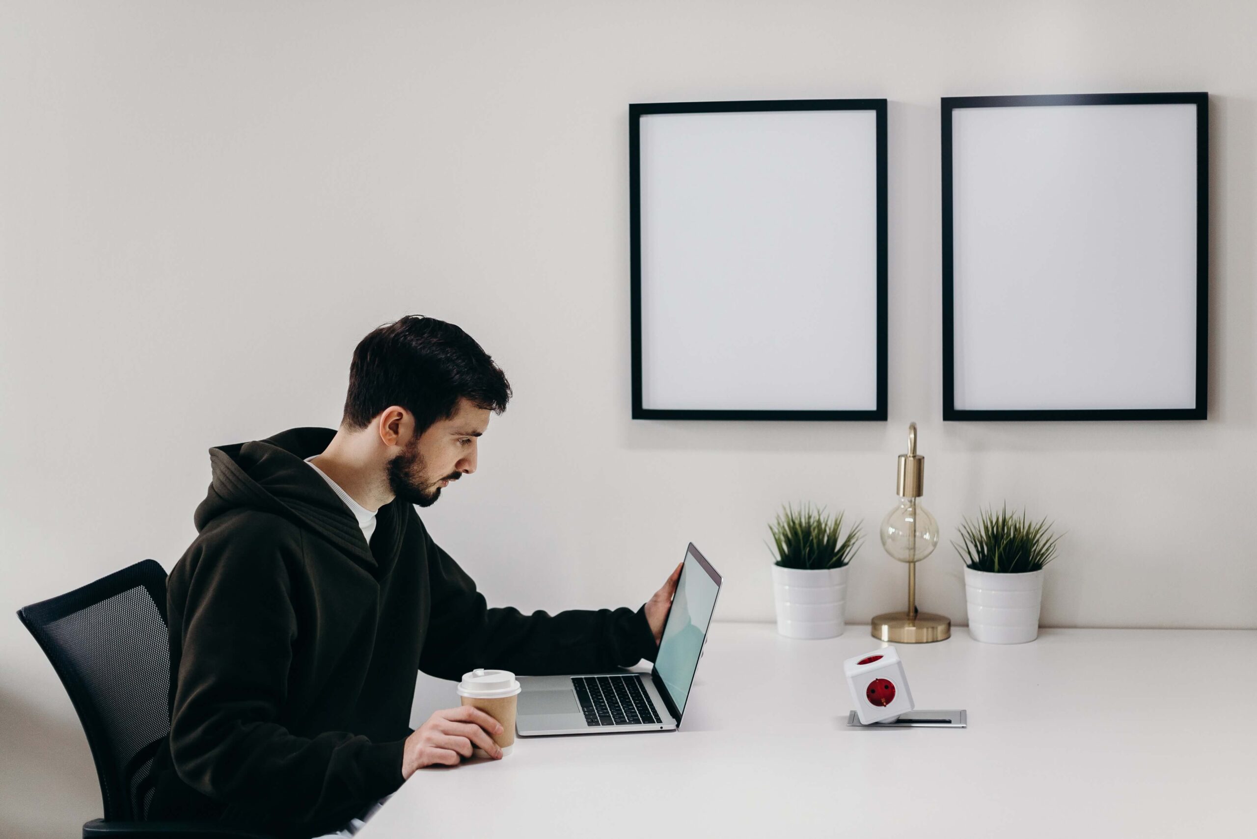 Man in a hoodie working on a laptop with coffee in a minimalist office setup, featuring plants and decor.