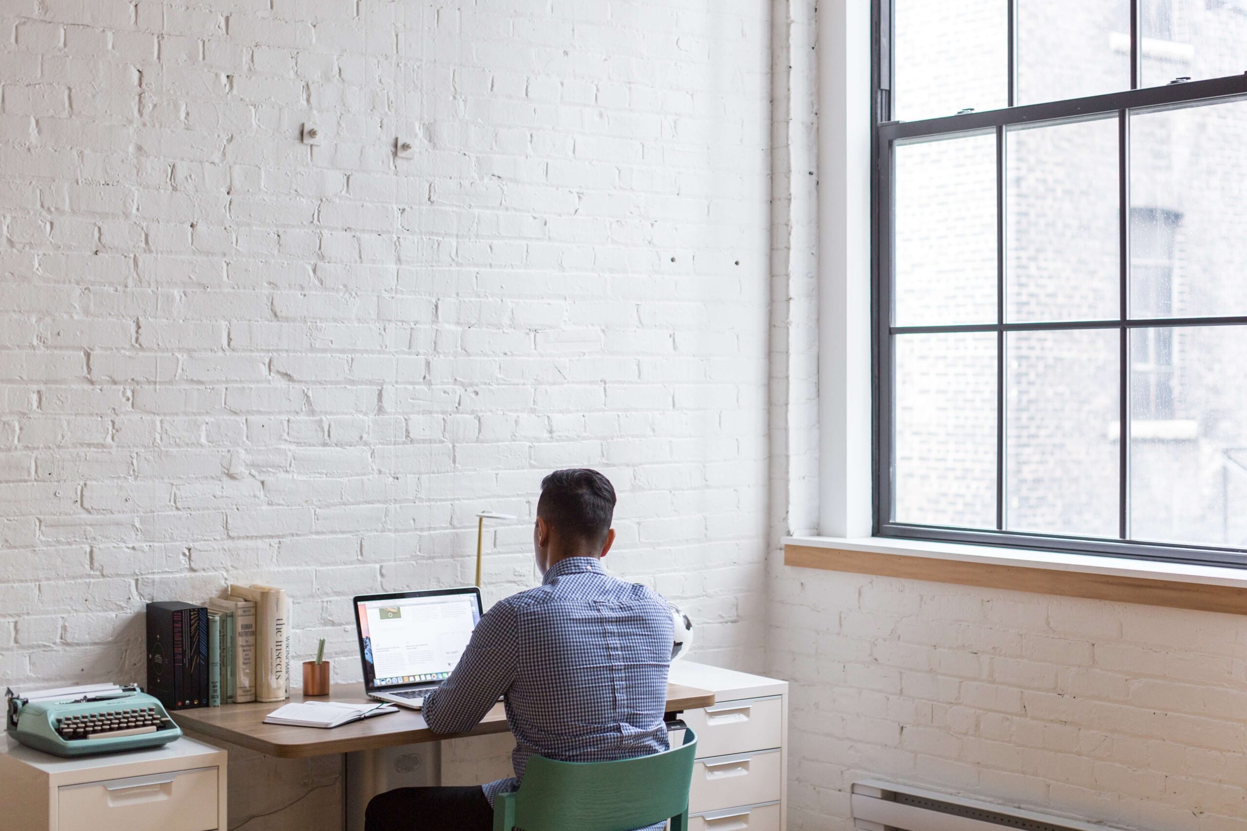 Man working on a laptop in a bright, minimalist home office with white brick walls and large windows.