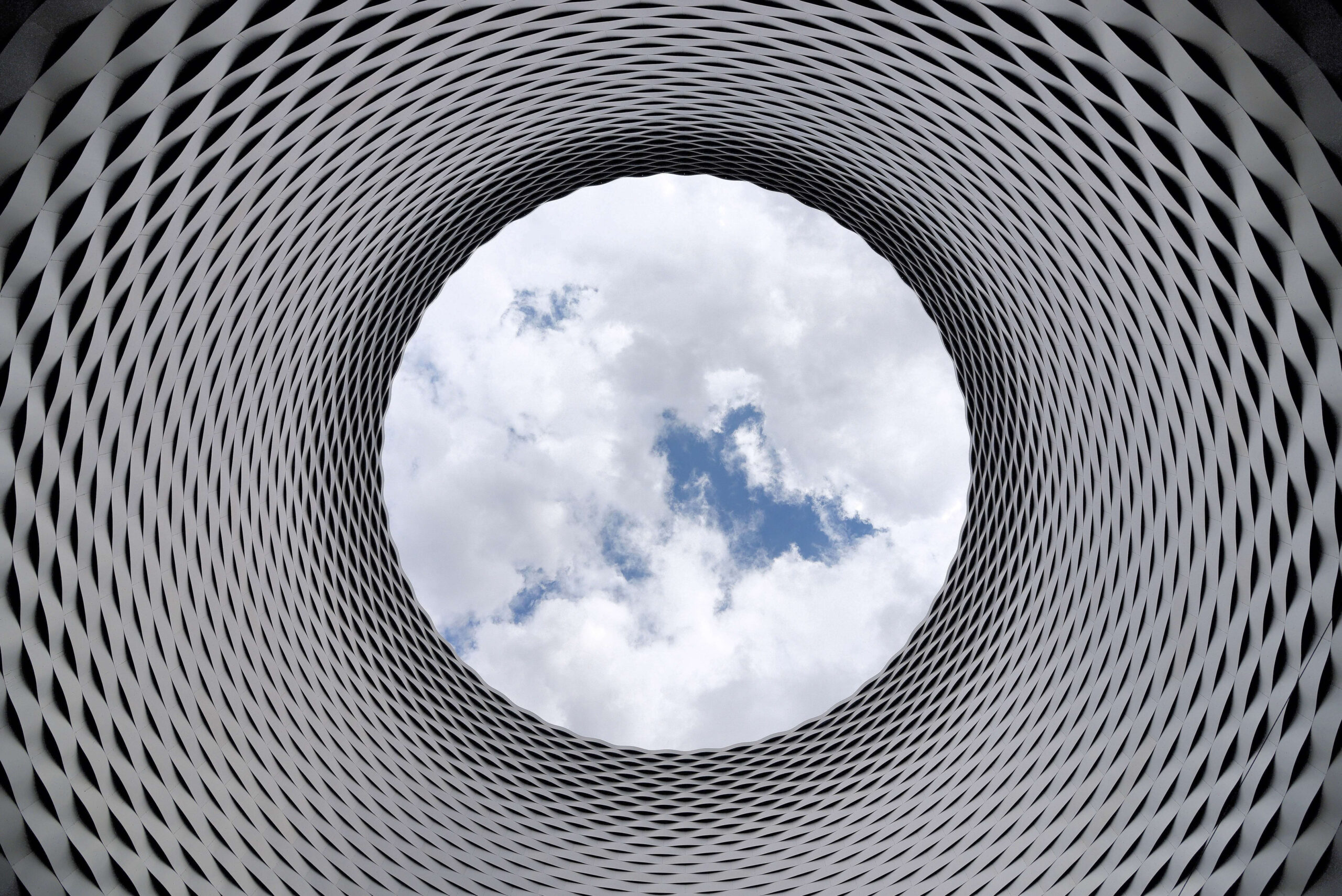 Futuristic architectural design with circular opening, looking up at cloudy sky through an intricate patterned structure.