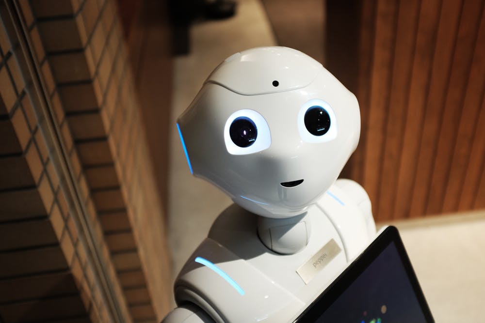 Friendly humanoid robot with glowing eyes standing indoors near a wooden wall, holding a tablet.