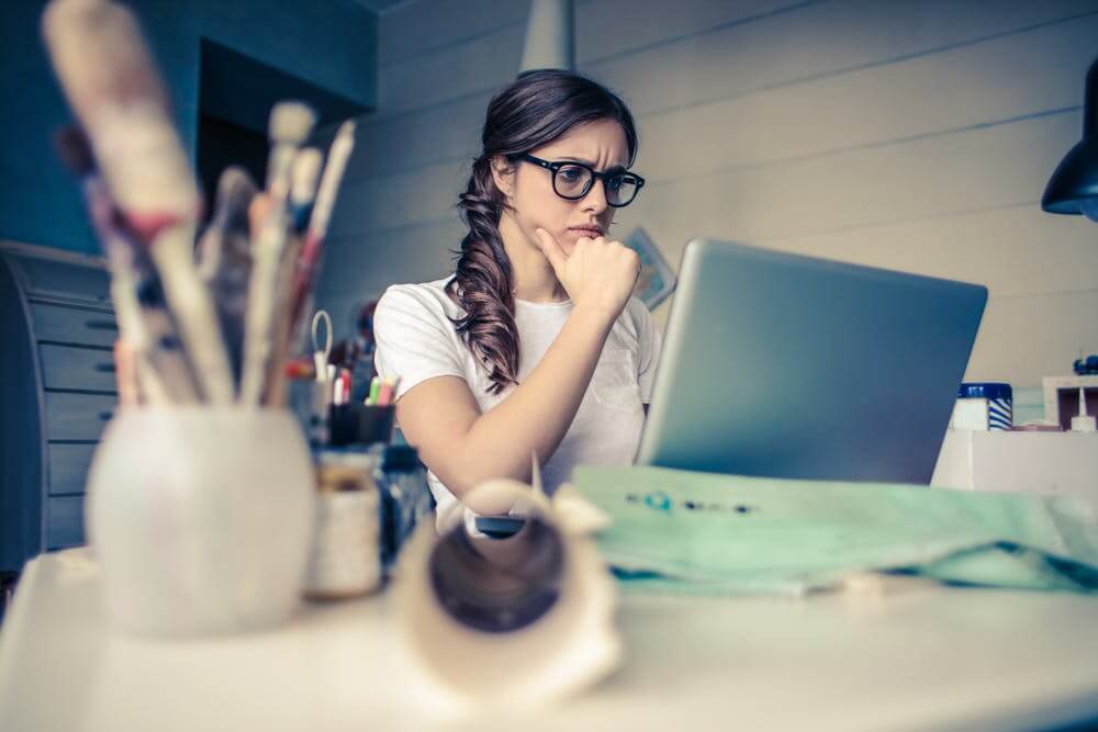 Woman focused on laptop at desk with paintbrushes, working creatively in a home studio environment.
