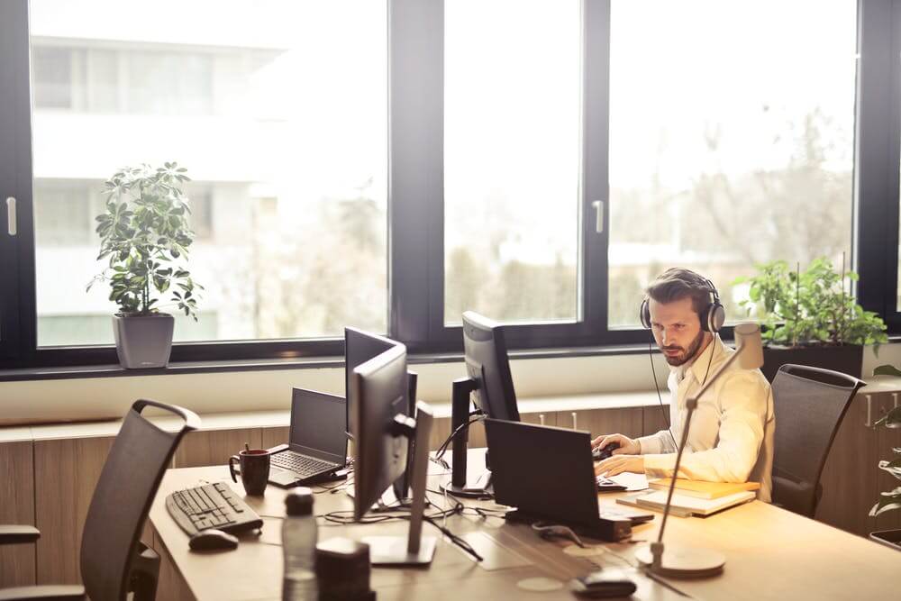 Man working at a desk with dual monitors and headphones, surrounded by plants, in a bright office setting.