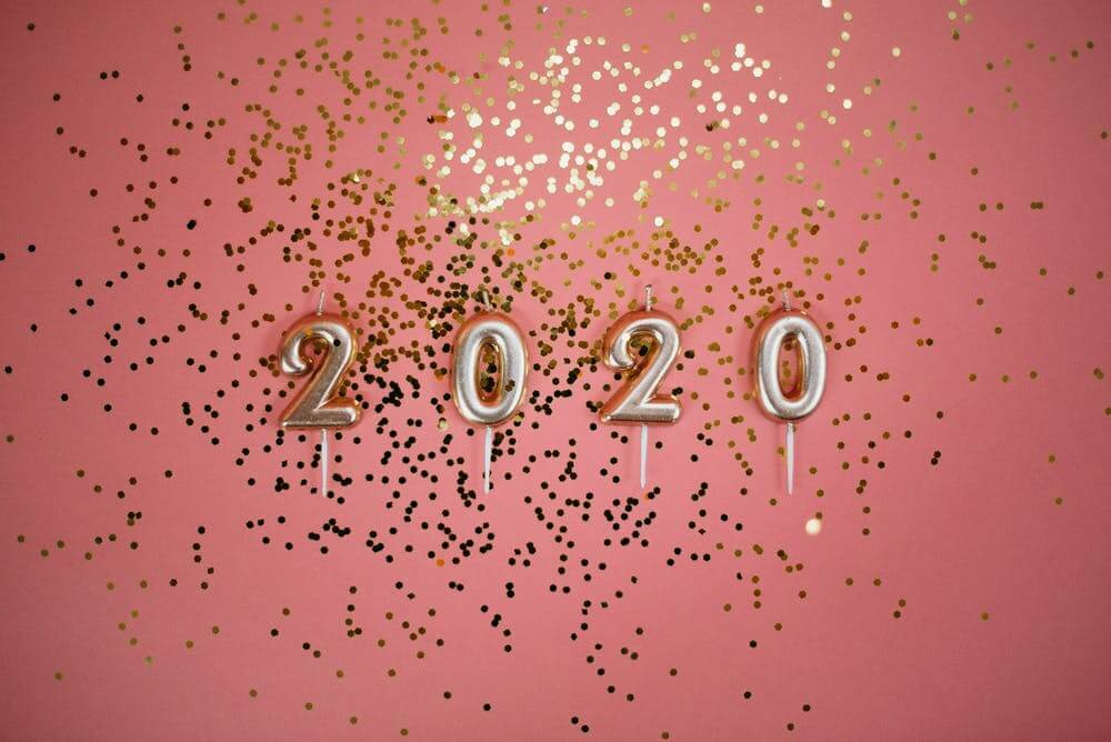 Silver 2020 candles on pink background with gold confetti for New Year celebration.