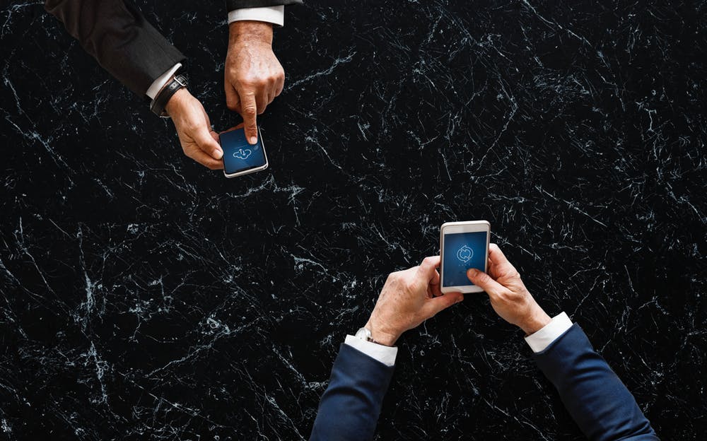 Two people in suits hold smartphones over a marble surface, both phones displaying cloud synchronization icons.