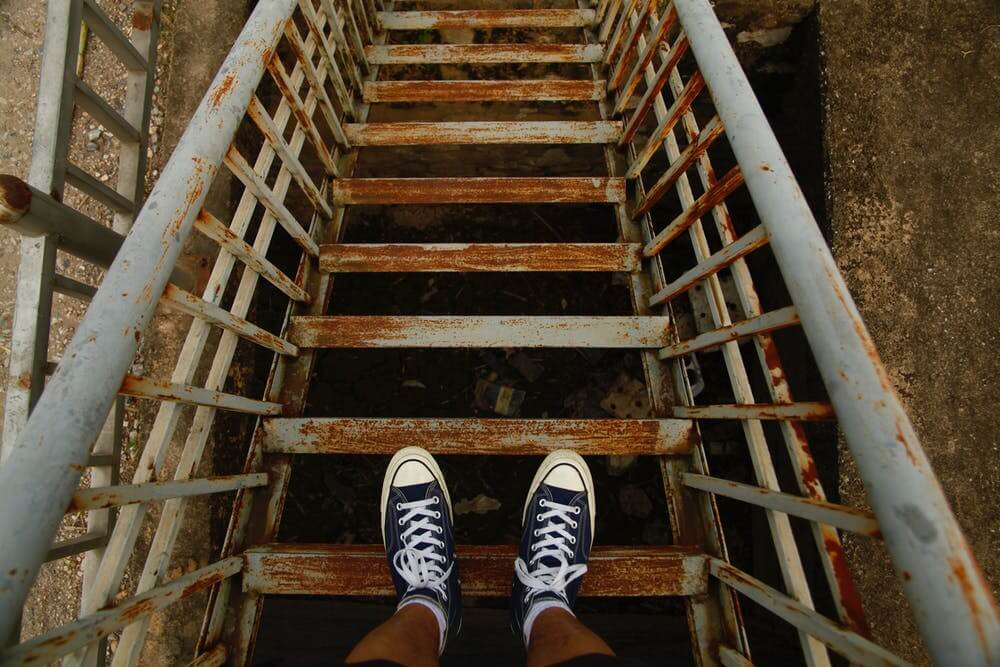 Looking down at feet in blue sneakers on old, rusty metal stairs, highlighting urban exploration and decay.