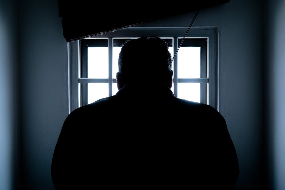 Silhouette of a person facing a barred window in a dimly lit room, suggesting confinement or introspection.