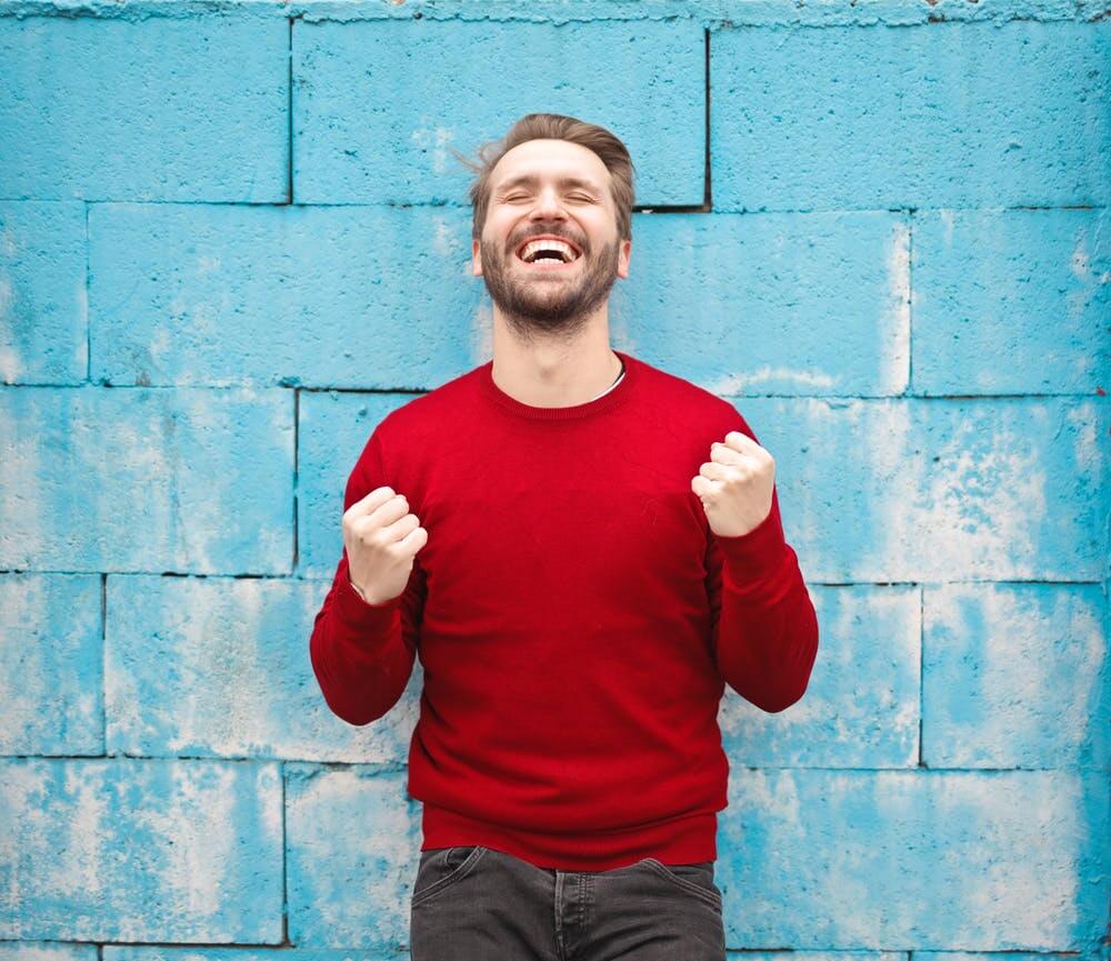 Man in red sweater celebrates joyfully against a blue brick wall background.