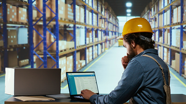Warehouse worker in a hard hat using a laptop for inventory management in a large storage facility.