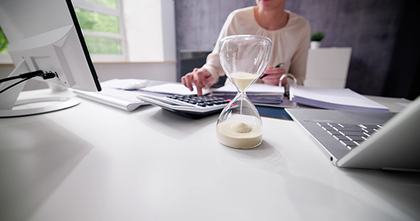 Hourglass on desk with woman calculating at computer, symbolizing time management and productivity in office setting.