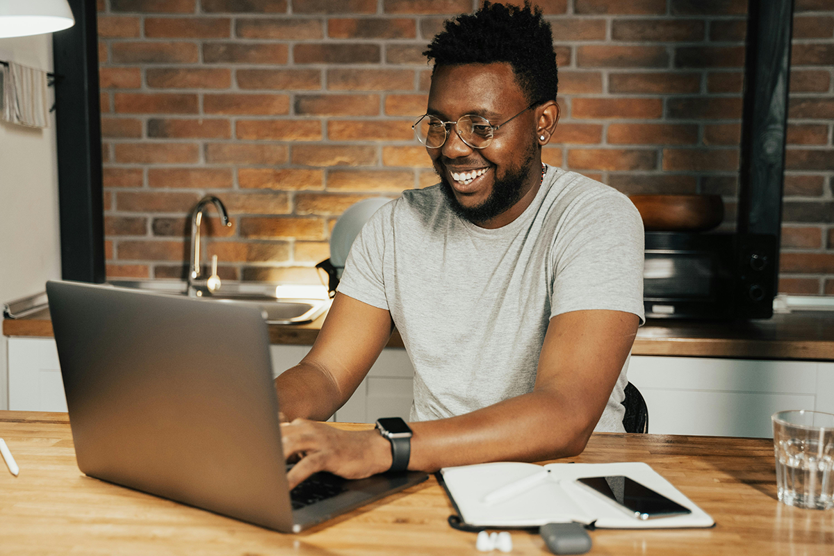 Man smiling while working on a laptop at a wooden table with notepad and phone, in a cozy kitchen setting.
