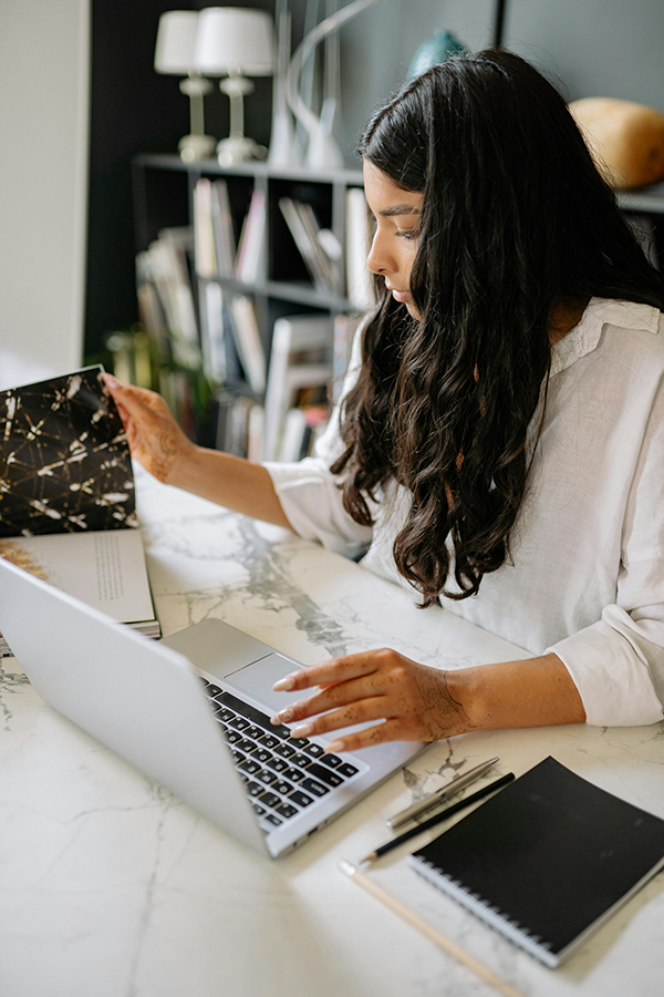 Woman working on laptop while reviewing a book at a marble desk with notebooks and pens.