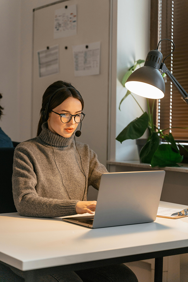 Woman with headset working on laptop at office desk, under lamp light.