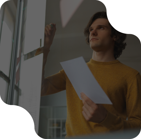 A person in a yellow sweater holds a paper and writes on a transparent board, focused and thoughtful, in an office setting.