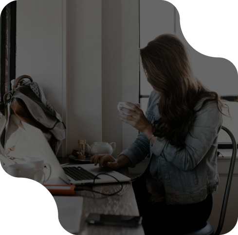 Woman in denim jacket working on laptop at a cafe, holding a coffee mug.