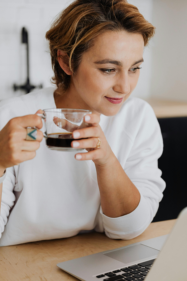 Person in a white shirt sips coffee while using a laptop at a wooden desk, smiling thoughtfully.