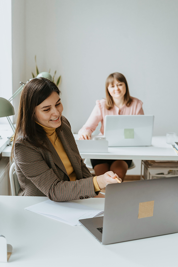Two women working at laptops in a bright office, one smiling and extending her arm.
