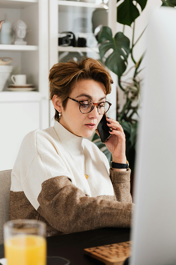 Woman in glasses working at a desk, talking on a phone with a computer screen and a glass of juice visible.
