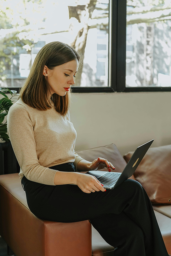 Woman working on laptop while seated on a sofa in a bright room, focused on screen.