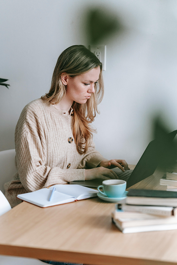 Woman working on laptop at a desk with a notebook and coffee, wearing a beige sweater, focused on her task.