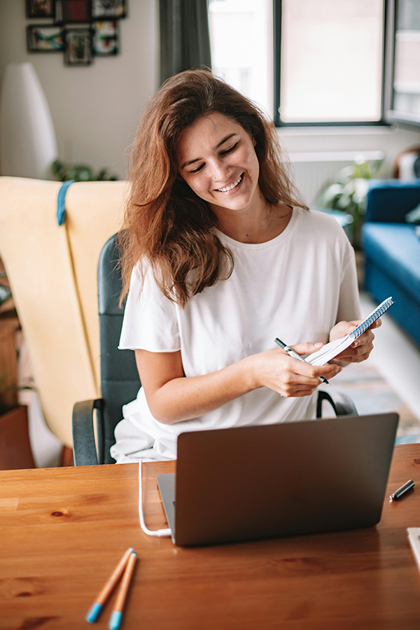 Smiling woman works on laptop, holding notepad in cozy home office setting.