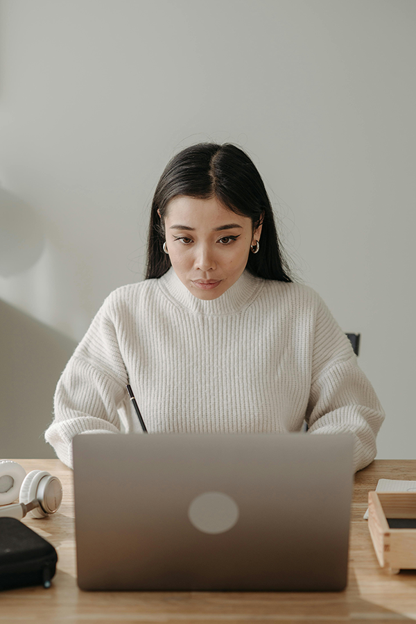 Woman focused on laptop work at wooden desk, wearing a white sweater, with headphones and a notebook beside her.