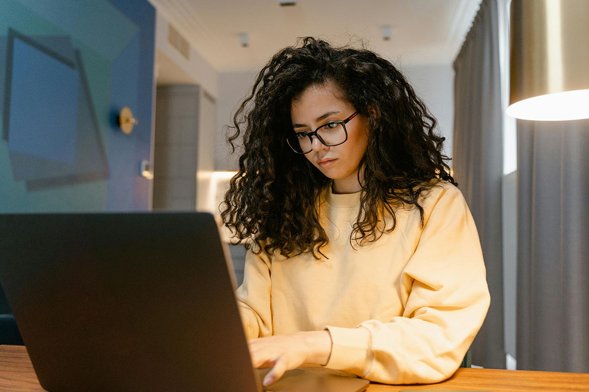 Woman in yellow sweater working on a laptop at a desk in a cozy room.