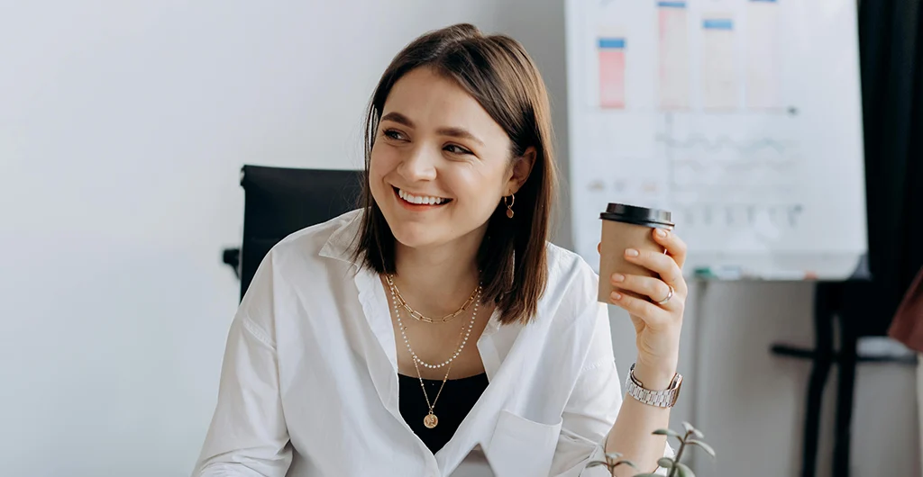 Smiling woman holding coffee at work desk with laptop and notepad, engaging in a friendly office meeting.
