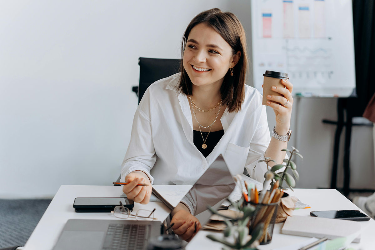 Smiling woman at desk with laptop, holding coffee and enjoying a relaxed work environment.
