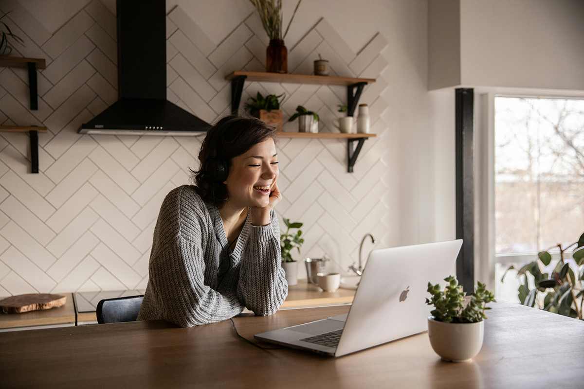 Woman smiling on video call while working from home in cozy kitchen setting with laptop and headphones.