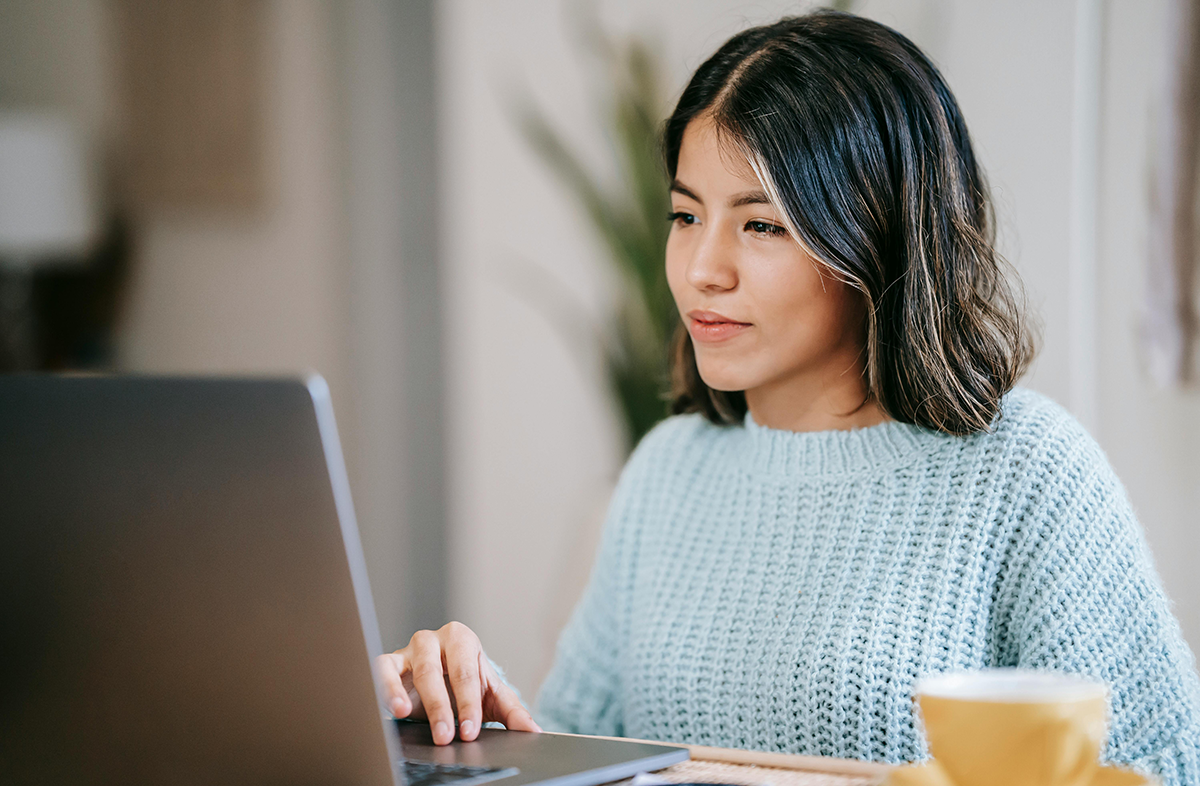 Woman in blue sweater using laptop at home, focused on screen, with coffee cup nearby.