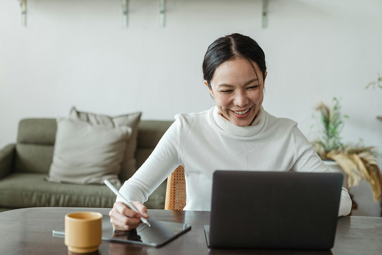 Smiling woman working on a laptop and tablet in a cozy home office with plants.