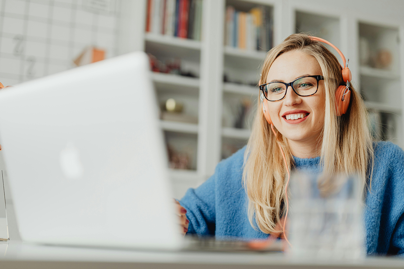 Smiling woman with glasses and headphones working on a laptop in a cozy home office.