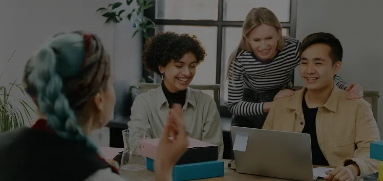 A diverse group of colleagues smiling and collaborating around a laptop in a modern office setting.