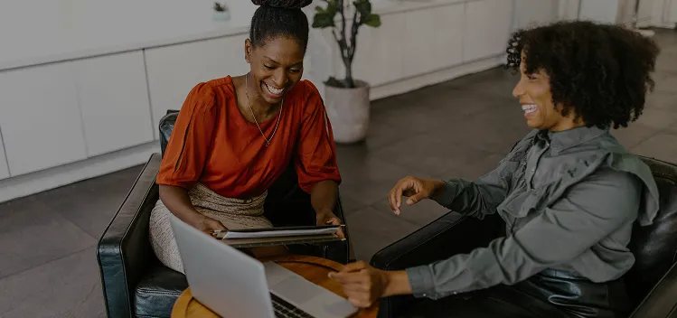 Two women smiling while working on a laptop in a modern office setting.