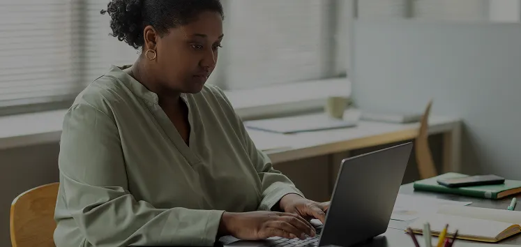 Woman in green blouse working on a laptop at a desk, focused in a bright office setting.