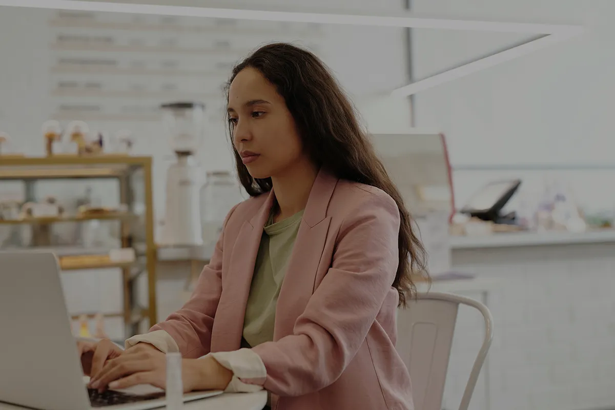 Woman in pink blazer working on a laptop in a bright, modern cafe setting.