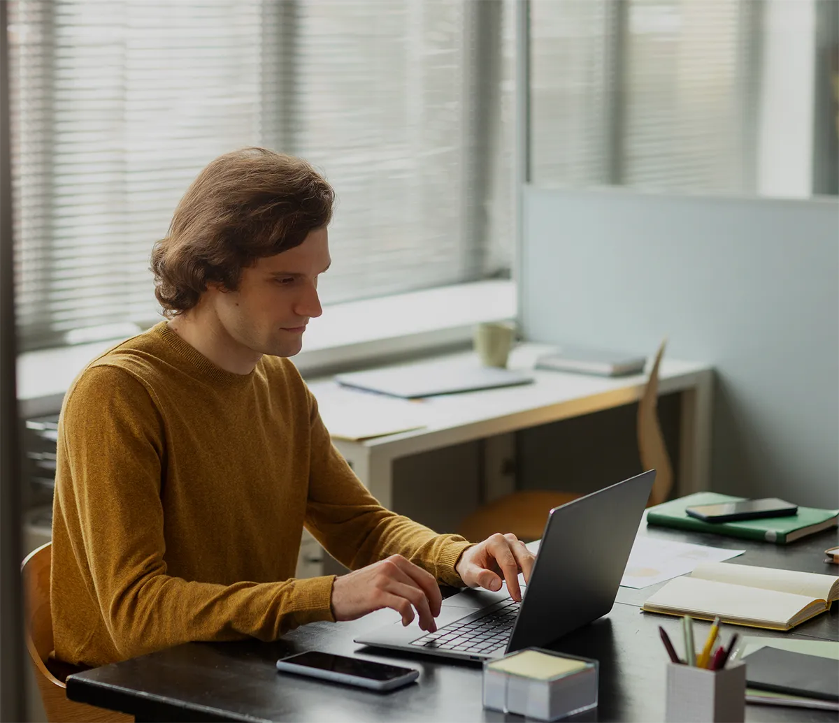 Person working on a laptop at an office desk, surrounded by notebooks and a smartphone, near a window with blinds.